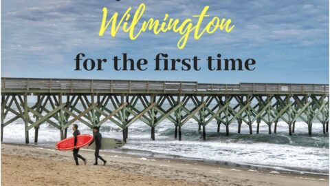 Two surfers walk on a sandy beach next to a wooden pier in Wilmington, NC.