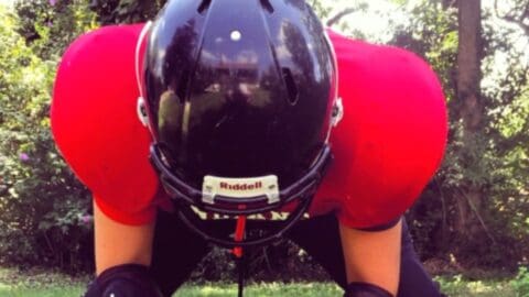 Football player in helmet and pads bends over on a grassy field.