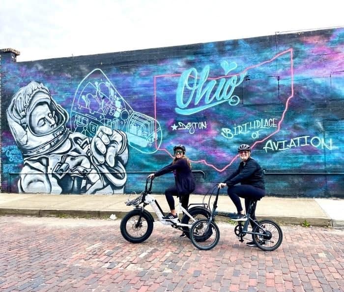 Adventure Mom and friend on e-bikes near aviation mural in Dayton
