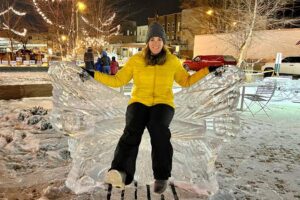 Woman in yellow jacket sits on ice butterfly sculpture in snowy outdoor setting.