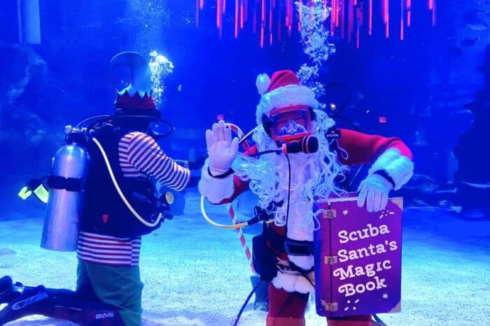 Scuba Santa with an elf underwater holding a magic book and waving.